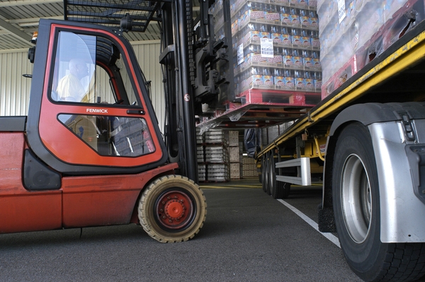 Un carrello elevatore, guidato da un operatore, sta caricando un pallet di bevande confezionate sul pianale di un camion